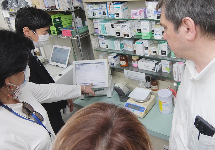 調剤室内で薬剤の計量機器について説明するスーツの男性とそれに耳を傾けるキルギスからの視察団