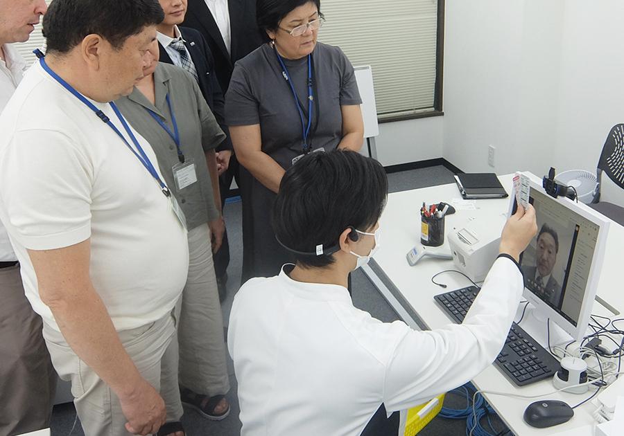 PCのカメラに向かってお薬を見せながらオンライン服薬指導のデモを行うケーシーを着た男性薬剤師とそれに耳を傾けるキルギスからの視察団